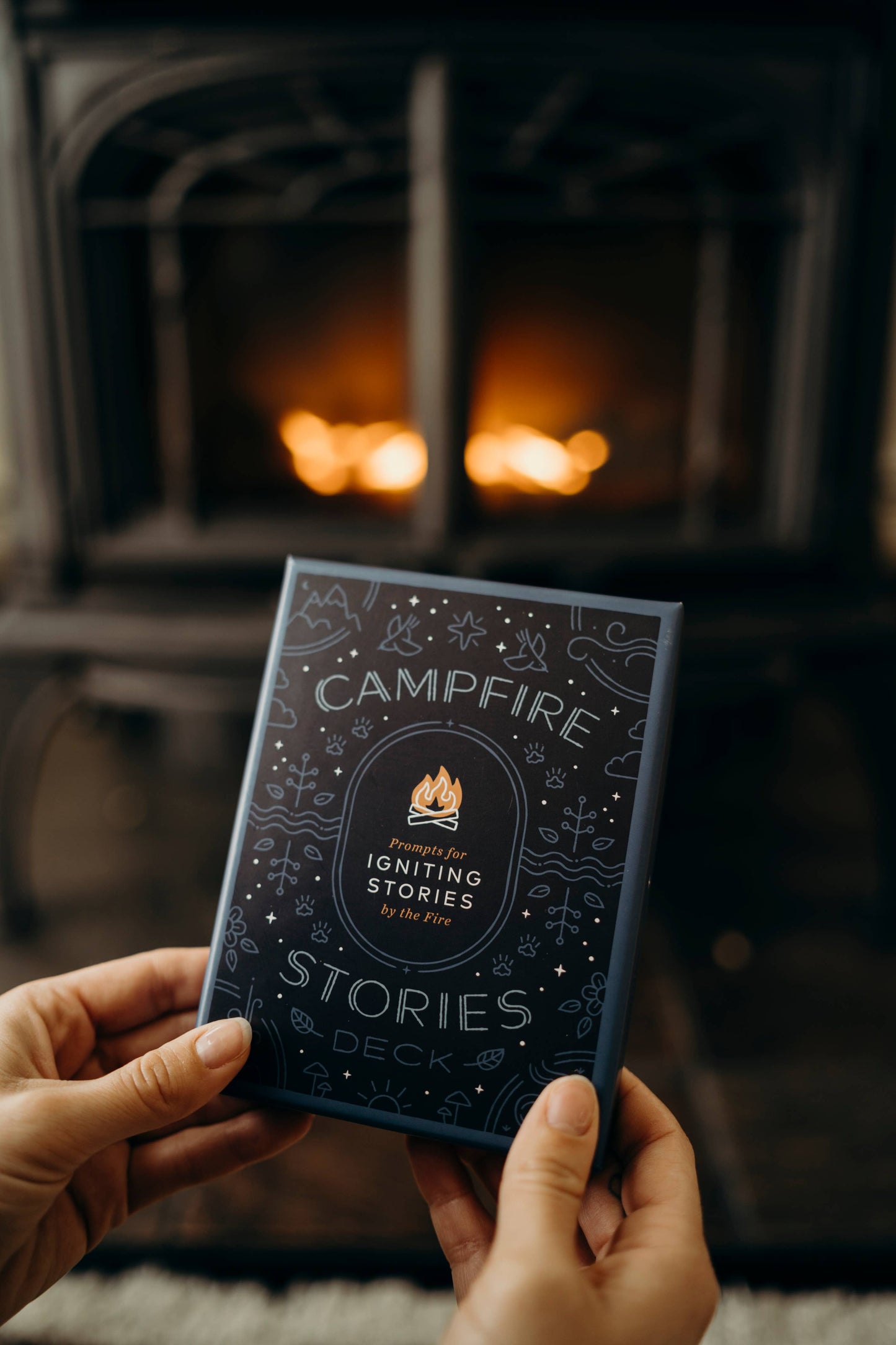 Person holding a 'Campfire Stories' card deck in front of a fireplace.