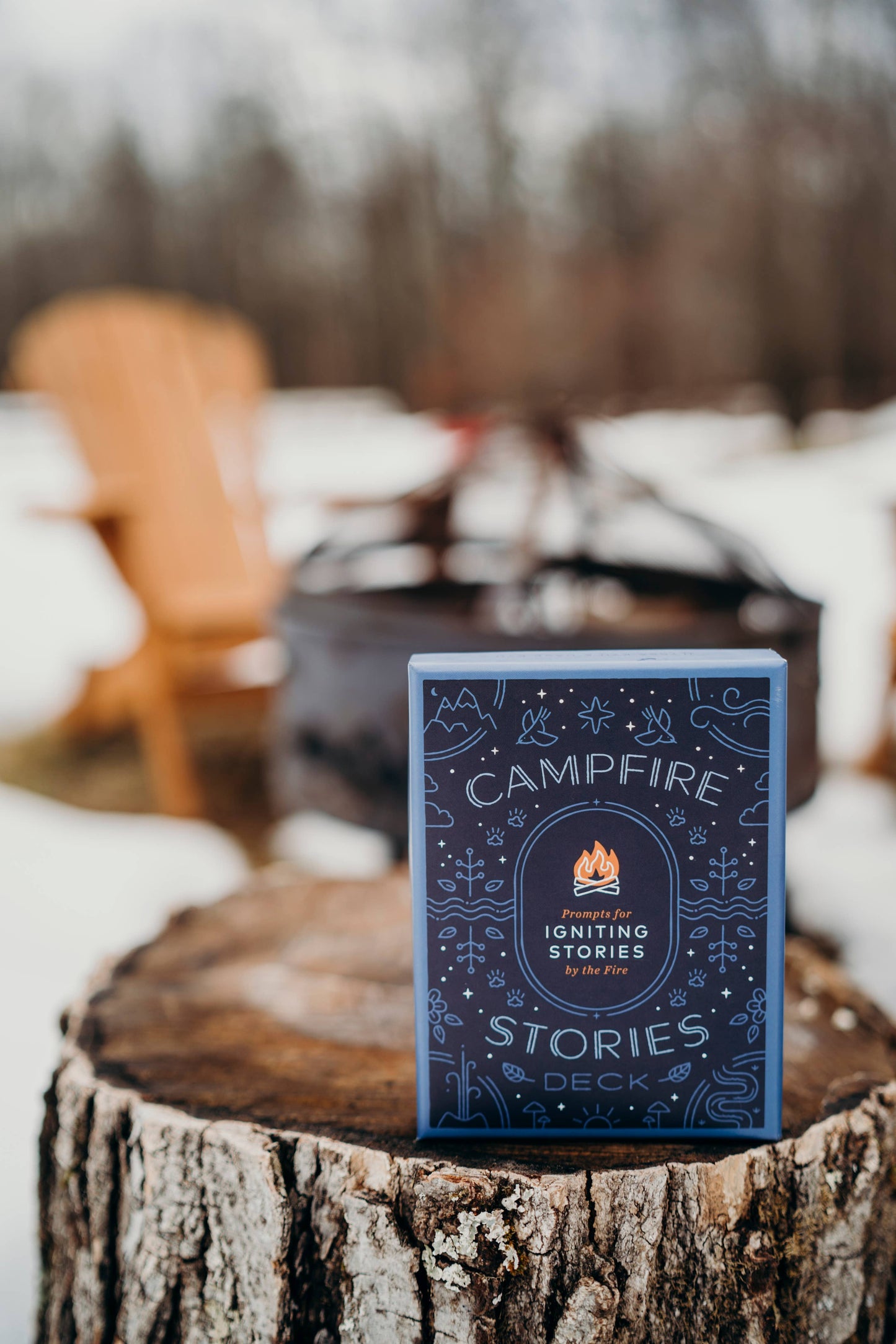 Card deck titled 'Campfire Stories' on a wooden stump with a blurred background of a fire pit and chairs.