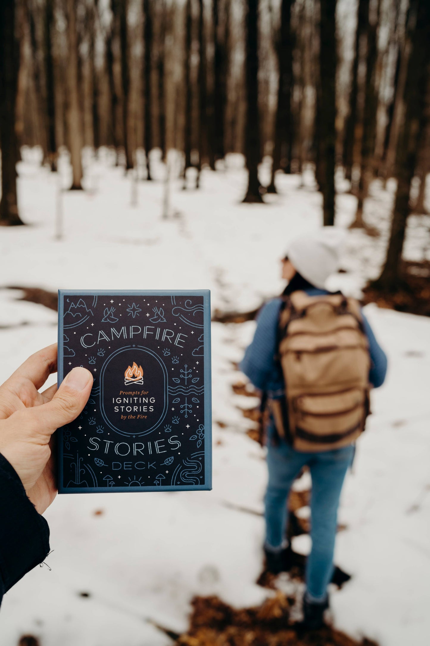 Person holding a book titled 'Campfire Stories' in a snowy forest with another person in the background.